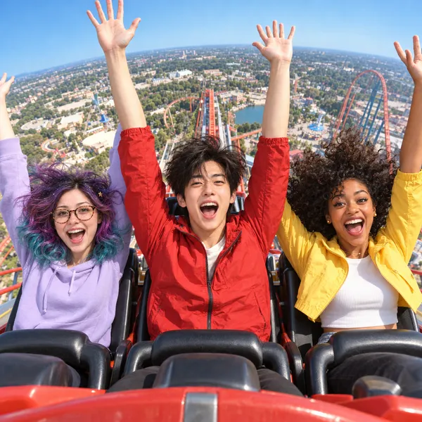Pip in a lilac hoodie and glasses, Jun in a red jacket and Maya in a yellow jacket and white top sit in a red roller coaster car, arms raised and mouths open, high above a theme park with coaster tracks and a lake under a blue sky.